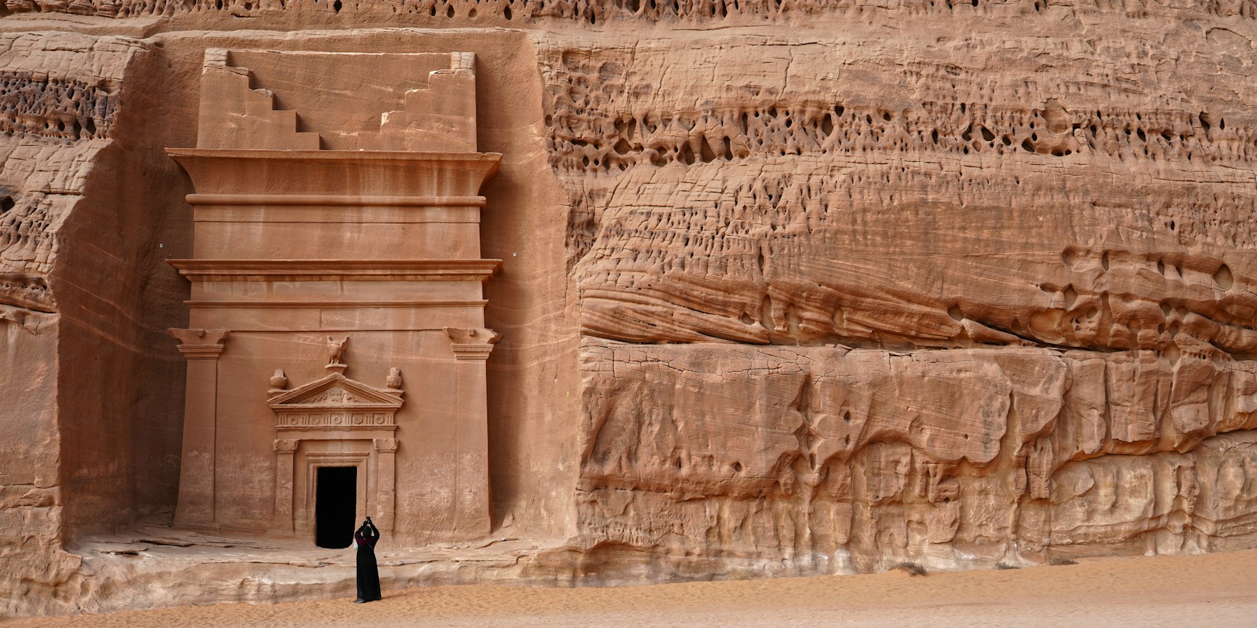 A visitor standing below an ancient sandstone tomb at Hegra